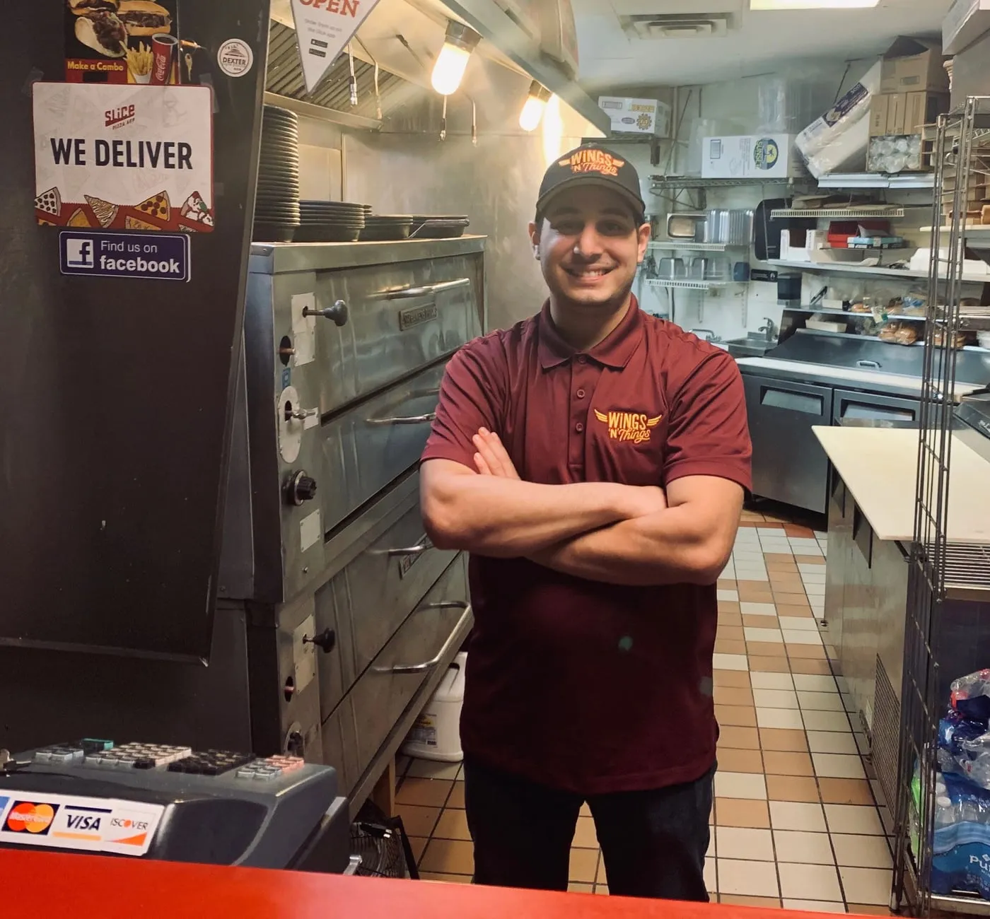 Team member in a Wings N Things shirt, arms crossed and smiling by the pizza ovens